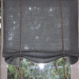 May include: A gray linen Roman shade with a frayed edge and a natural rope cord. The shade is partially lowered, revealing a view of trees outside the window. The rope cord has a decorative tassel.