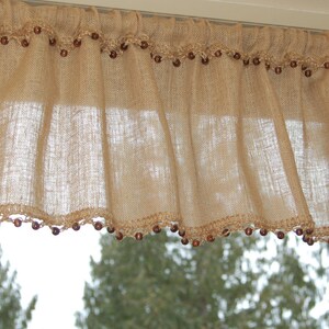 May include: A burlap valance with a crocheted trim and brown beads. The valance is hanging in a window with a view of trees outside.