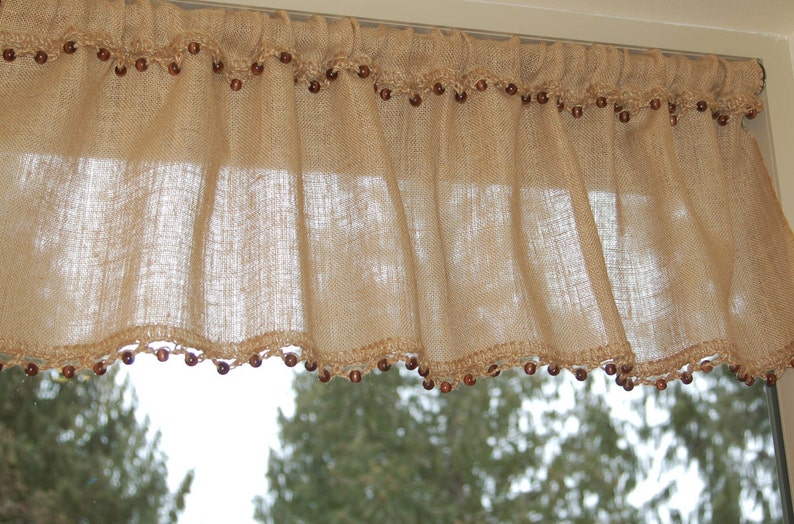 May include: A burlap valance with a crocheted trim and brown beads. The valance is hanging in a window with a view of trees outside.
