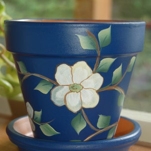 May include: A blue painted terracotta flower pot with a white flower and green leaves painted on the side. The pot has a matching saucer.
