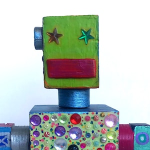 May include: A colourful wooden robot toy with a green head, blue body, and multi-coloured arms and legs. The robot is decorated with rhinestones and painted with geometric patterns.