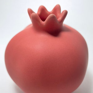 May include: A pink ceramic vase shaped like a pomegranate. The vase has a smooth, matte finish and a slightly textured top.