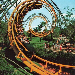 May include: A vintage postcard image of a roller coaster with yellow tracks and green supports, looping through the air. Passengers are visible in the cars, enjoying the ride. Lush green trees and a blue sky provide the backdrop.