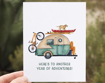 May include: A watercolor illustration of a vintage camper van with a bicycle, a canoe, and a dog on top. The camper is green and white with a wood paneling design. The text "Here's to another year of adventures!" is written below the camper.