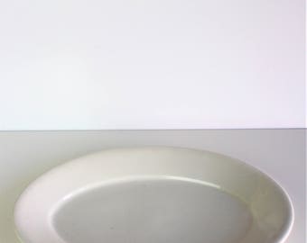 May include: A white, oval-shaped serving platter. The ceramic dish has a smooth surface and a slightly raised edge. The platter is empty and sits on a white surface, suitable for serving food.