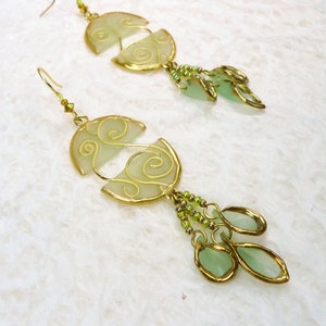 May include: A pair of gold earrings with green glass teardrop shapes and a swirl design. The earrings are made of metal and have a delicate, feminine look.