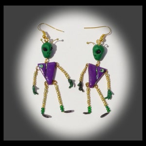 Puede incluir: Un par de pendientes colgantes con figuras de extraterrestres verdes con detalles en morado y dorado. Los extraterrestres tienen cuerpos de cuentas y brazos y piernas dorados.