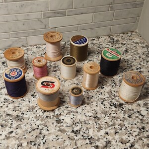 Puede incluir: Una colección de once carretes de hilo de coser de madera vintage en varios colores, incluyendo blanco, negro, rosa, gris, verde oliva y azul marino. Cada carrete presenta una etiqueta de papel circular en la parte superior con marcas como Star y Blossom.