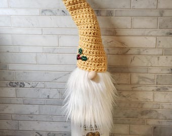 May include: A decorative bottle gnome made to fit over a bottle. The gnome has a long, curved, mustard-yellow crocheted hat with a holly accent. It features a white faux fur beard and a wooden nose. The bottle reads "150 Years Sparkling Cider".
