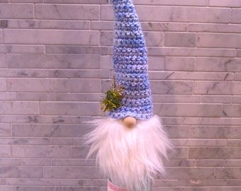 May include: A whimsical gnome decoration crafted from a repurposed wine bottle. The gnome features a long, blue and white crocheted hat, a fluffy white beard, and a wooden nose. A small sprig of greenery adorns the hat. The bottle is clear, with a pink label.