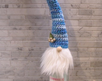 May include: A decorative gnome wine bottle topper. The gnome has a long, pointed blue and white crocheted hat, a fluffy white beard, and a wooden nose. A small gold embellishment is on the hat. The topper is designed to fit over a wine bottle.