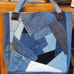 May include: A patchwork denim tote bag with blue jean fabric pieces in various shades of blue and gray. The bag has denim handles and brown leather accents. The bag is rectangular and appears to be handmade.