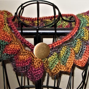 Puede incluir: Un calentador de cuello de ganchillo multicolor con cierre de botón de latón. El calentador de cuello se muestra en un maniquí negro.