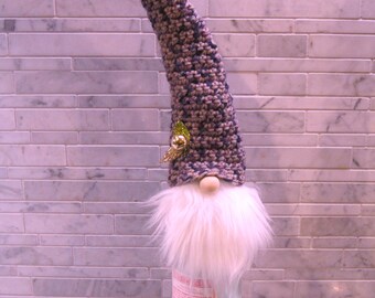 May include: A decorative gnome bottle topper. The gnome has a long, pointed hat in shades of blue and tan, a fluffy white beard, and a wooden nose. A small gold leaf and bead adorn the hat. The gnome sits atop a clear glass bottle.