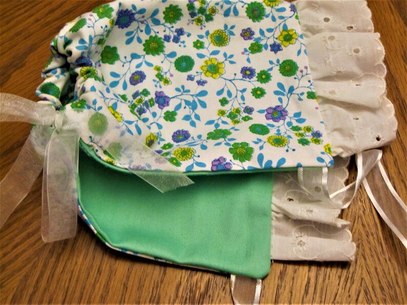 Puede incluir: Una bolsa de tela verde y blanca con estampado floral y ribete de encaje blanco. La bolsa tiene un cierre de cord&oacute;n y una cinta blanca atada alrededor.