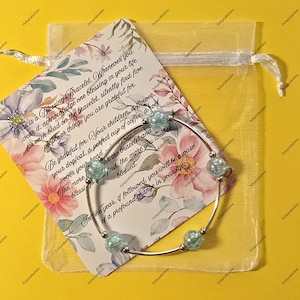 May include: A silver bracelet with four light blue, speckled beads, displayed with a card and a sheer yellow gift bag. The card has floral designs and text that reads "This is a Blessing Bracelet."