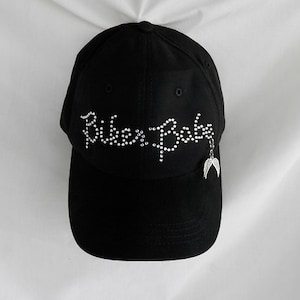 May include: Black baseball cap with the words "Ride or Die" in rhinestones. A silver charm with angel wings hangs from the brim.