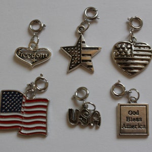 May include: Six silver charm pendants with patriotic themes. The charms include a heart with the word "Freedom" on it, a star with red and white stripes, a heart with the American flag, a small American flag, the word "USA", and a square with the words "God Bless America".