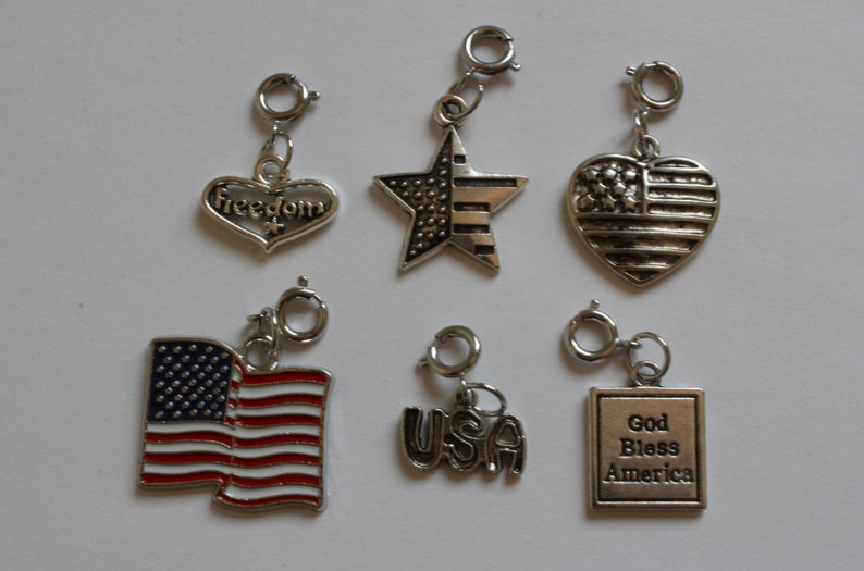 May include: Six silver charm pendants with patriotic themes. The charms include a heart with the word "Freedom" on it, a star with red and white stripes, a heart with the American flag, a small American flag, the word "USA", and a square with the words "God Bless America".