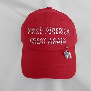 Pu&ograve; includere: Un cappellino da baseball rosso con la scritta "MAKE AMERICA GREAT AGAIN" in strass. Un piccolo ciondolo a forma di bandiera americana pende dalla visiera.