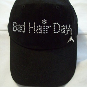 May include: Black baseball cap with the words "Bad Hair Day" spelled out in rhinestones. A small silver hair dryer charm hangs from the side of the cap.