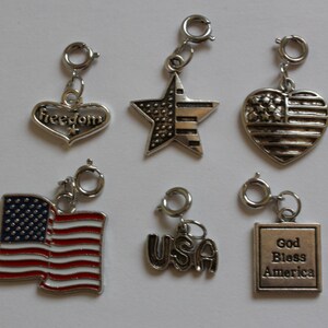 May include: Six silver charm pendants with patriotic themes. The charms include a heart with the word "Freedom" on it, a star with red and white stripes, a heart with the American flag, a small American flag, the word "USA", and a square with the words "God Bless America".