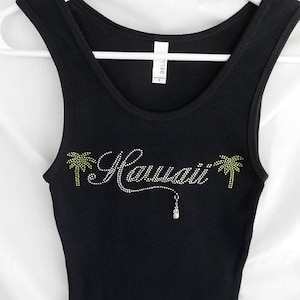 May include: Black tank top with a rhinestone design that says "Hawaii" with palm trees on either side and a pineapple charm hanging from the word.