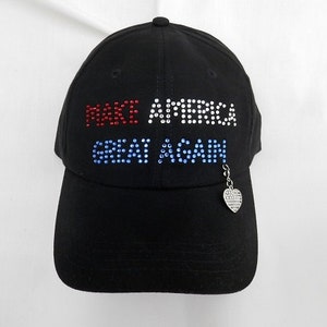 May include: Black baseball cap with the words "Make America Great Again" in rhinestones. The rhinestones are red, white, and blue. A small silver charm in the shape of a heart with an American flag design hangs from the cap.