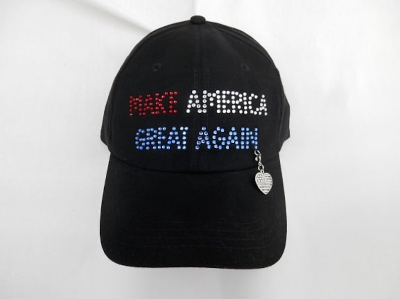 May include: Black baseball cap with the words "Make America Great Again" in rhinestones. The rhinestones are red, white, and blue. A small silver charm in the shape of a heart with an American flag design hangs from the cap.