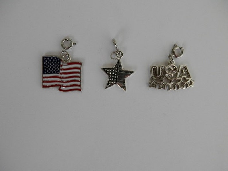 May include: Three silver charm pendants with patriotic themes. The first charm is a red, white and blue American flag. The second charm is a silver star with a blue and white striped design. The third charm is a silver pendant with the word "USA" in raised letters and five small stars.