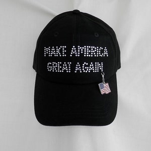 May include: A black baseball cap with the phrase "Make America Great Again" in white rhinestones. A small American flag charm hangs from the brim.