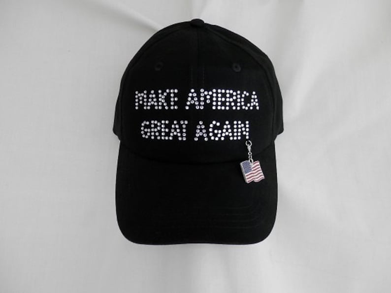 May include: A black baseball cap with the phrase "Make America Great Again" in white rhinestones. A small American flag charm hangs from the brim.