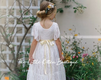 May include: A young girl wearing a white lace dress with a yellow sash and a flower crown. The dress has a tiered skirt and short sleeves. The girl is standing in a garden with green plants and flowers.