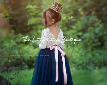 Op de afbeelding: Een jong meisje draagt een witte kanten jurk met een donkerblauwe tule rok en een roze strik. Ze draagt een gouden kroon en staat in een grasachtig gebied. De tekst "The Little Kitten Boutique" is zichtbaar in de afbeelding.