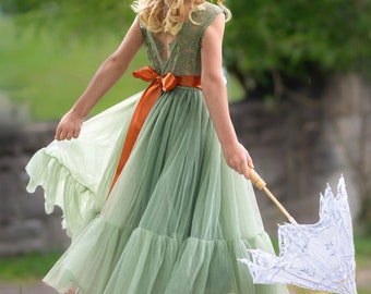 May include: A young girl wearing a sage green tulle dress with a ruffled hem and an orange satin sash. She is holding a white lace parasol and has her hair styled in loose curls.