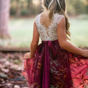 Könnte beinhalten: Burgunderfarbenes Kleid mit weißem Spitzenoberteil und floral besticktem Rock. Das Kleid hat eine Samtschärpe und ist für besondere Anlässe für Kinder geeignet. Der Rock ist tiefrot.