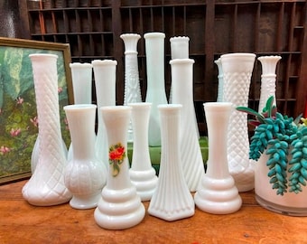 May include: A collection of 11 white milk glass bud vases with various designs and textures. The vases are arranged on a wooden surface.