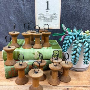 May include: A collection of vintage wooden thread spool holders with black wire loops. A calendar page from July 1948 is in the background. The spools are arranged on top of antique green books, with a succulent plant in a white pot.