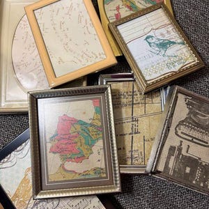 May include: A collection of framed art pieces with various designs. The frames are in gold, silver, and black. The art includes maps, musical notes, and a bird illustration. The frames are rectangular and oval.