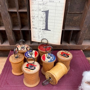 May include: Eight wooden spools with wire loops, each spool has a different label, some labels have red, white, and blue colors, the labels say "Liberty" and "40". The spools are sitting on a burgundy book with gold lettering.
