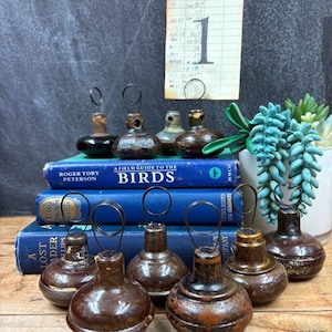 May include: A collection of dark brown, rounded wooden objects with metal wire loops on top, arranged on three stacked blue books. A vintage library card with the text "Heiner, Lynn" and "Hardworking Puppies" is held upright by one of the wire loops.