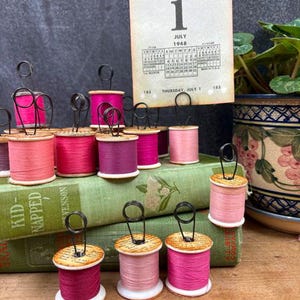 May include: A collection of vintage wooden thread spools in various shades of pink and magenta, each topped with a black wire coil holder. A rectangular card displaying the calendar date Thursday, July 1, 1948, is held upright by one spool.