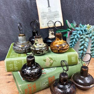 May include: A collection of vintage metal photo holders with a circular base and wire loop. The holders are in various colors, including silver, black, and brown, and are arranged on top of antique green books. A calendar page is in the background.