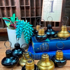 May include: A collection of eight vintage glass and metal table number holders. The holders are black and gold with a wire loop at the top. They are arranged on a wooden surface with a stack of books in the background.