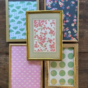 May include: Five rectangular picture frames with metallic gold-toned borders. The frames contain various decorative papers featuring patterns of green umbrellas, pink and blue flowers, pink horses, red floral sprigs, and green polka dots.
