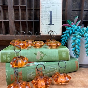 May include: Nine orange glass place card holders with wire rings. The holders are arranged on top of two green books. The books have titles that include "Kid-Napped" and "The Trail".