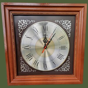 May include: A square wall clock with a brown wooden frame. The clock face is silver with black Roman numerals and black hands, with a red second hand. The clock has a dark brown background with white decorative accents. The brand name "Sunbeam" is visible.