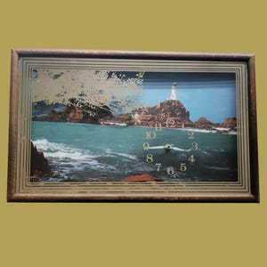 May include: A rectangular wall clock featuring a landscape print of a lighthouse on a rocky coast with turquoise water. The clock face is integrated into the image with gold-colored numbers and white hands, all encased in a dark brown frame.