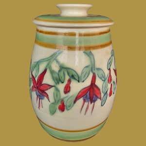 May include: A ceramic jar with a lid, featuring a cream-colored base decorated with hand-painted fuchsia flowers in shades of red and purple, green leaves, and horizontal bands of light green and brown encircling the top and bottom.