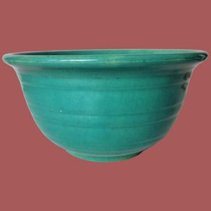 May include: A teal ceramic bowl with a wide rim and a rounded body. The bowl features horizontal ridges around its exterior, adding texture to the smooth, glossy surface. The bowl is set against a solid, reddish-brown background.